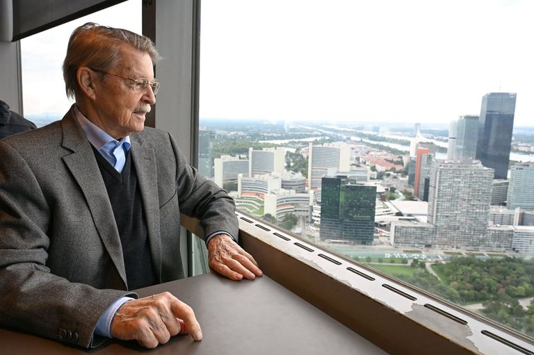 Ein älterer Mann in Anzug und Brille sitzt in einem Bürohochhaus an einem Schreibtisch und blickt aus dem Fenster auf eine Stadtlandschaft mit Gebäuden, einem Fluss und einer Brücke.
