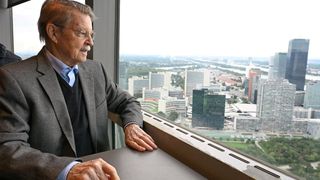 Ein älterer Mann in Anzug und Brille sitzt in einem Bürohochhaus an einem Schreibtisch und blickt aus dem Fenster auf eine Stadtlandschaft mit Gebäuden, einem Fluss und einer Brücke.