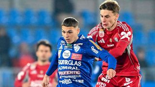 Zwei Fußballspieler auf einem Spielfeld, einer in einem blauen Trikot und der andere in einem roten Trikot, kämpfen um den Ball.