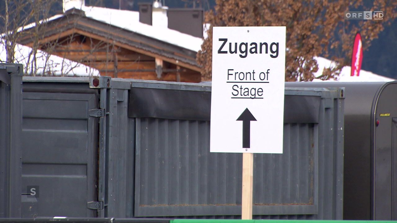 Ein weißes Schild mit schwarzem Text ist auf einem Metallzaun platziert. Es steht 'Zugang Front of Stage' mit einem Pfeil nach oben. Das Schild befindet sich wahrscheinlich am Eingang zur Bühne.