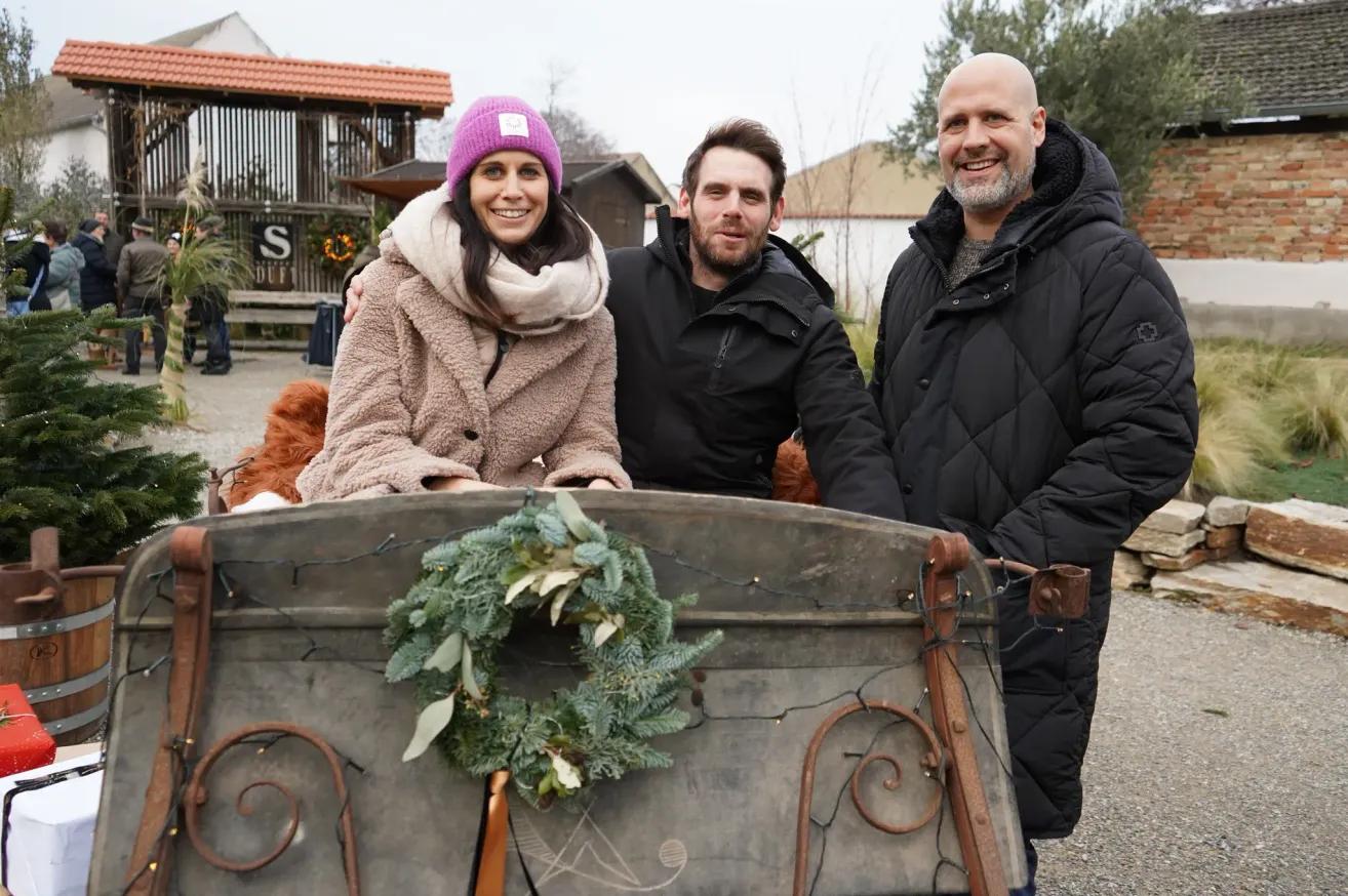 Drei Menschen lächeln und stehen vor einem Schlitten mit einem Kranz darauf. Sie sind warm gekleidet. Dahinter stehen Gebäude und Bäume.