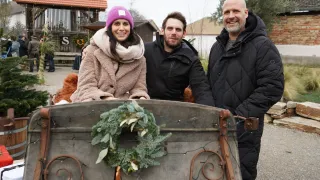 Drei Menschen lächeln und stehen vor einem Schlitten mit einem Kranz darauf. Sie sind warm gekleidet. Dahinter stehen Gebäude und Bäume.