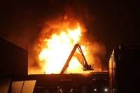 Ein großes Feuer umgibt ein Gebäude in der Nacht. Eine Feuerwehrleiter ist auf die Flammen ausgerichtet, möglicherweise um das Feuer zu löschen. Das Gebäude ist teilweise sichtbar mit eingeschalteten Lichtern.