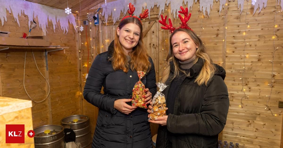Two women wearing Christmas headbands and black coats stand in a festive room with wooden walls and decorations. They hold bags of snacks.