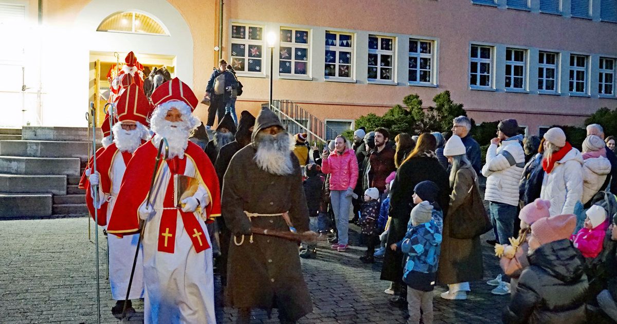 Zwei Menschen in Santa-Kleidung gehen vor einer Menschenmenge, einer in einem roten Gewand und der andere in einem braunen. Dahinter steht eine vielfältige Gruppe von Menschen, einschließlich Kindern, vor einem Gebäude mit Weihnachtsthema.