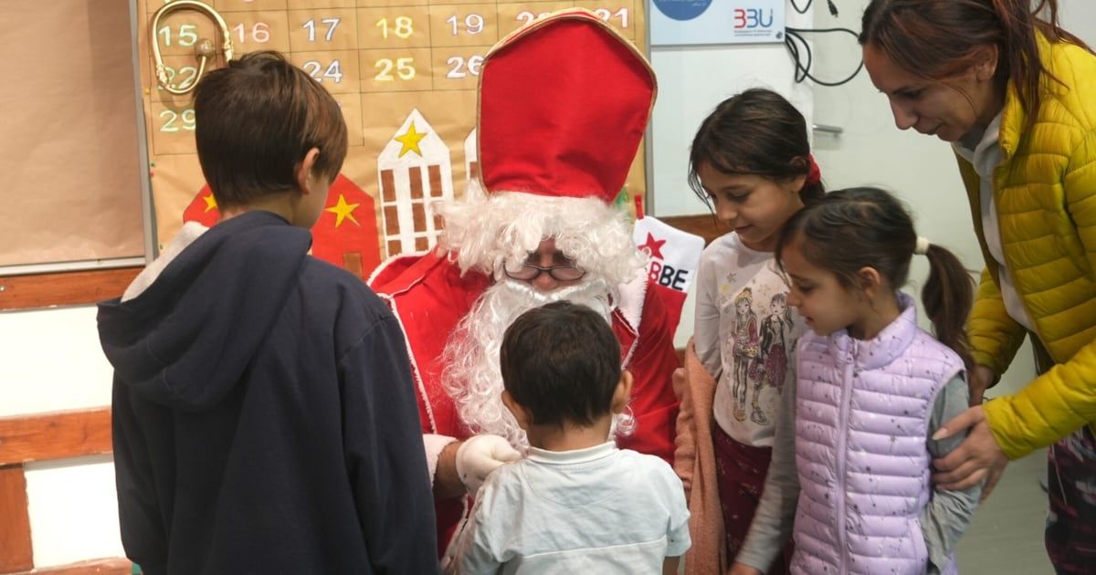 A man dressed as Santa Claus talks to children in a classroom setting, surrounded by a festive calendar backdrop.