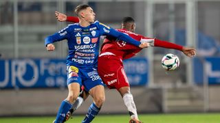 Zwei Fußballspieler in blauen und roten Trikots sind in einem Zweikampf. Der Spieler in Blau trägt ein Trikot mit mehreren Sponsorenlogos. Der Spieler in Rot verteidigt den Ball.