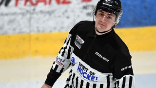 Ein Eishockeyspieler ist auf dem Eisfeld und trägt ein schwarz-weiß gestreiftes Schiedsrichtertrikot, einen schwarzen Helm und klare Brille. Das Trikot hat die Aufschrift Spirit of Hockey auf der Vorderseite.
