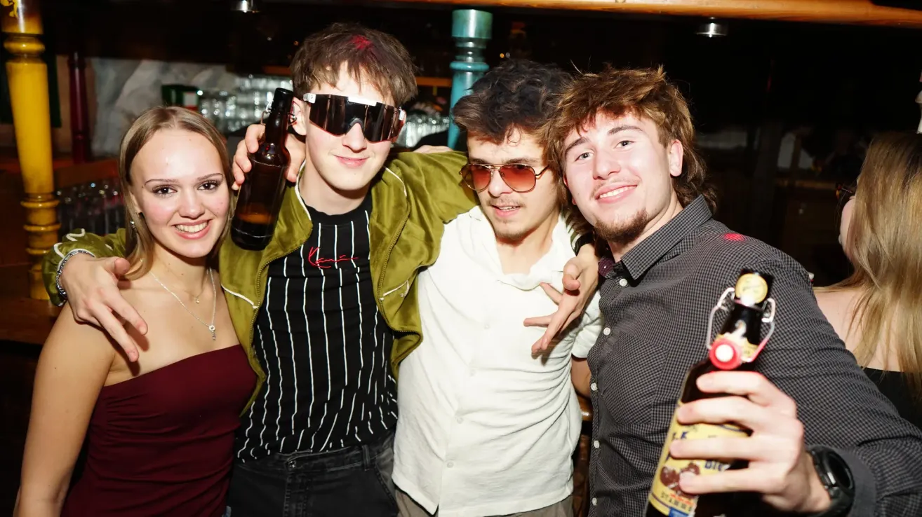 Vier junge Erwachsene posieren für ein Foto in einer Bar, einer hält eine Bierflasche.