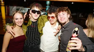 Vier junge Erwachsene posieren für ein Foto in einer Bar, einer hält eine Bierflasche.