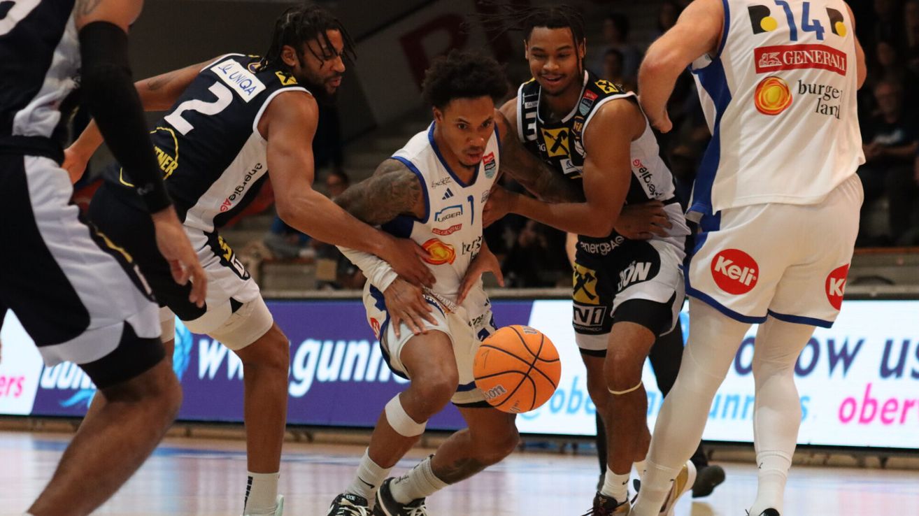 Basketballspieler im Spiel, einer dribbelt den Ball, zwei andere versuchen, ihn zu blocken, mit Publikum im Hintergrund.