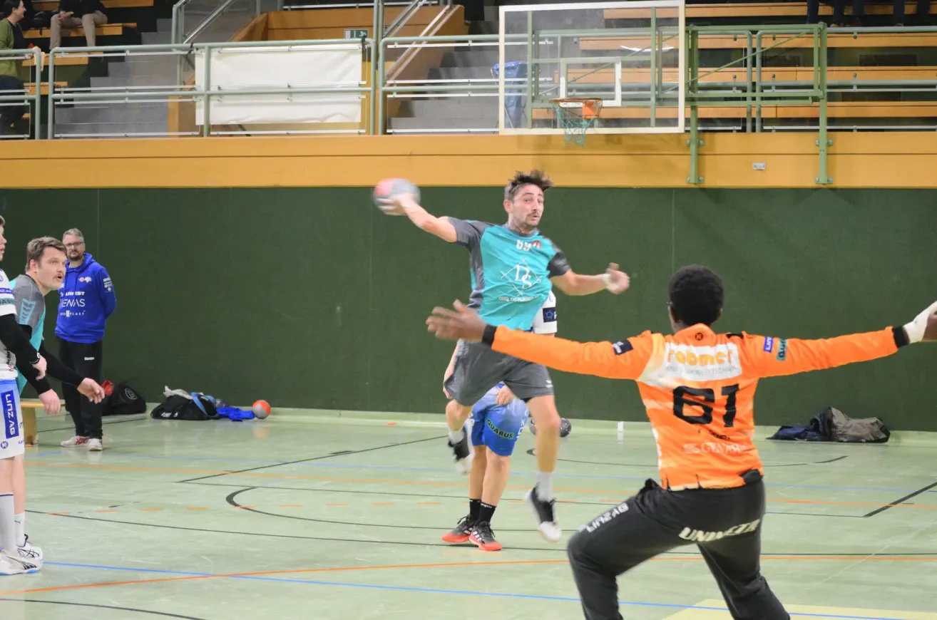 Hallenhandballspiel. Ein Spieler in einem türkisfarbenen Trikot wirft den Ball. Ein anderer Spieler in Orange verteidigt. Der Platz hat blaue Markierungen.