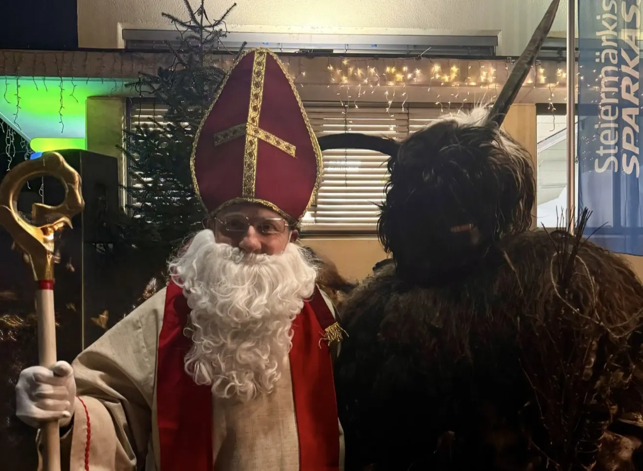 Ein Mann in einer Weihnachtsmann-Verkleidung steht neben einer weiteren Person in einem gehörnten Kostüm und lächelt in die Kamera. Dahinter steht ein Weihnachtsbaum und Lichterketten.