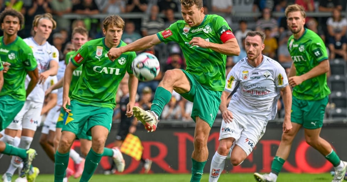 Drei Fußballspieler auf dem Spielfeld, einer tritt den Ball, ein anderer verteidigt und einer nähert sich, mit Zuschauern im Hintergrund.