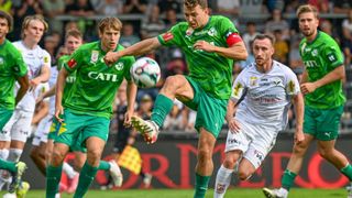 Drei Fußballspieler auf dem Spielfeld, einer tritt den Ball, ein anderer verteidigt und einer nähert sich, mit Zuschauern im Hintergrund.