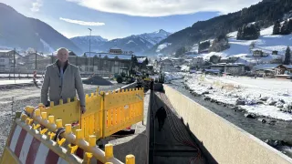 Ein Mann steht auf einer Brücke in einer verschneiten Landschaft, mit Bergen und einem Fluss im Hintergrund. In der Nähe befinden sich Baumaschinen und eine gelbe Absperrung.