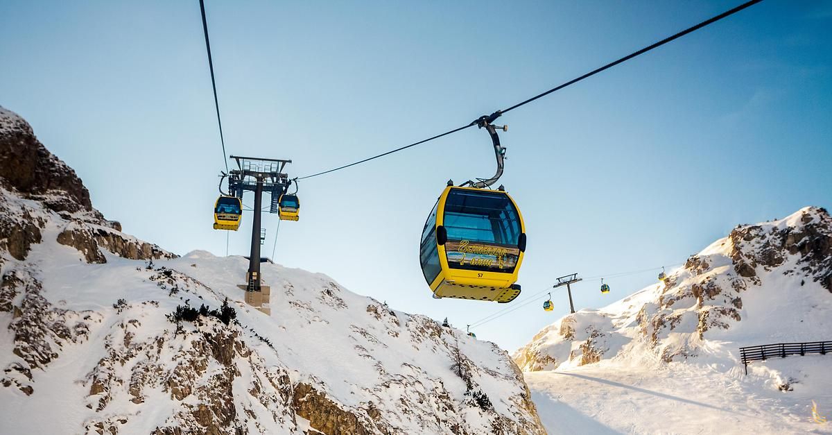 Eine gelbe Seilbahn schwebt in der Luft und durchquert einen verschneiten Berg, während eine weitere Seilbahn im Hintergrund absteigt.
