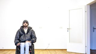 Ein Mann im Rollstuhl, der eine Mütze und eine Jacke trägt, sitzt in einem Raum mit weißer Wand, Tür und Steckdose. Er schaut nach vorne, hinter ihm befindet sich eine helle Wand und ein Holzboden.