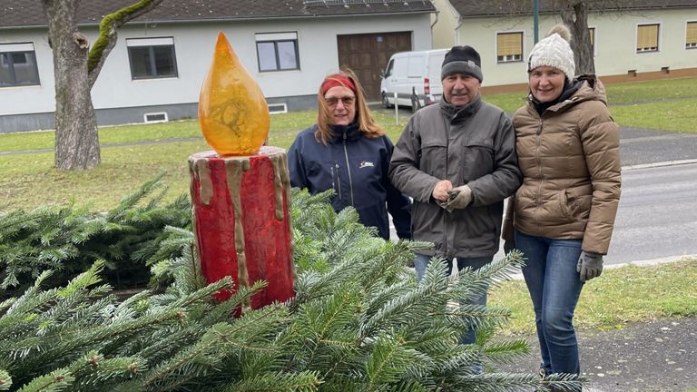 Drei Menschen stehen draußen neben einer großen Kerzenskulptur. Die Frau hat eine rote Kopfband, und der Mann trägt eine Mütze. Sie lächeln.