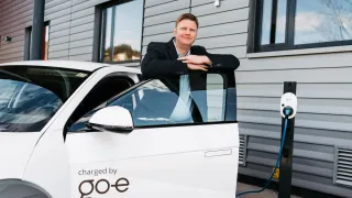 Ein Mann steht neben einem weißen Elektroauto mit offener Tür und lehnt sich mit einem Lächeln daran. Das Auto hat eine Ladestation und ein Schild mit der Aufschrift geladen von go-e.