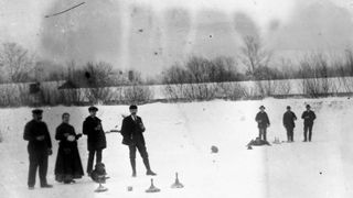 Eine Gruppe von Menschen in Winterkleidung spielt ein Spiel auf einem verschneiten Feld. Ein Hund liegt in der Nähe, und Spielteile sind auf dem Boden verstreut.