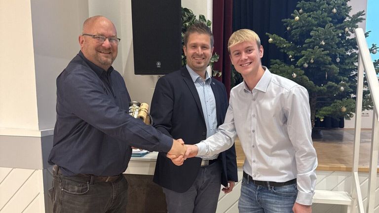 Drei Männer in einem Raum, zwei Hände schütteln. Der linke Mann hält eine Trophäe, der rechte in einem blauen Hemd lächelt. Dahinter ein Weihnachtsbaum und ein Lautsprecher.