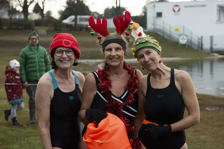 Drei Frauen in Badeanzügen und Weihnachtskopfbändern lächeln für die Kamera. Die Frau in der Mitte hält eine Tasche, und der Hintergrund zeigt einen Rasenbereich mit einem Gebäude und Bäumen.