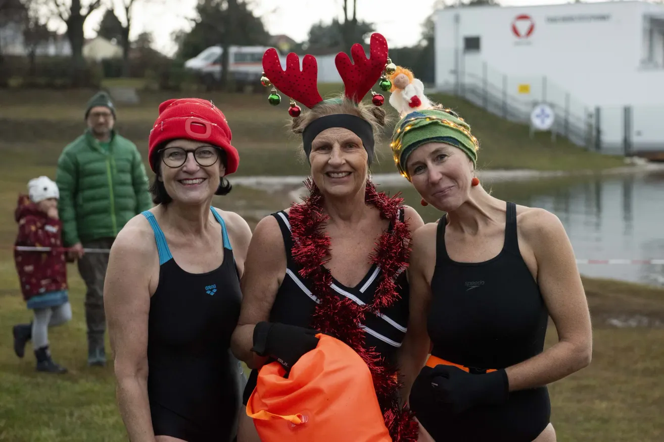 Drei Frauen in Badeanzügen und Weihnachtskopfbändern lächeln für die Kamera. Die Frau in der Mitte hält eine Tasche, und der Hintergrund zeigt einen Rasenbereich mit einem Gebäude und Bäumen.