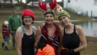 Drei Frauen in Badeanzügen und Weihnachtskopfbändern lächeln für die Kamera. Die Frau in der Mitte hält eine Tasche, und der Hintergrund zeigt einen Rasenbereich mit einem Gebäude und Bäumen.