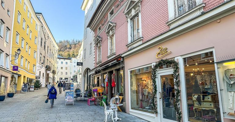 Ein Fußgänger läuft auf einer Kopfsteinpflasterstraße an Geschäften vorbei. Rechts zeigt ein Geschäft Artikel an und ist mit Grünpflanzen dekoriert. Im Hintergrund sind Gebäude und ein klarer blauer Himmel zu sehen.