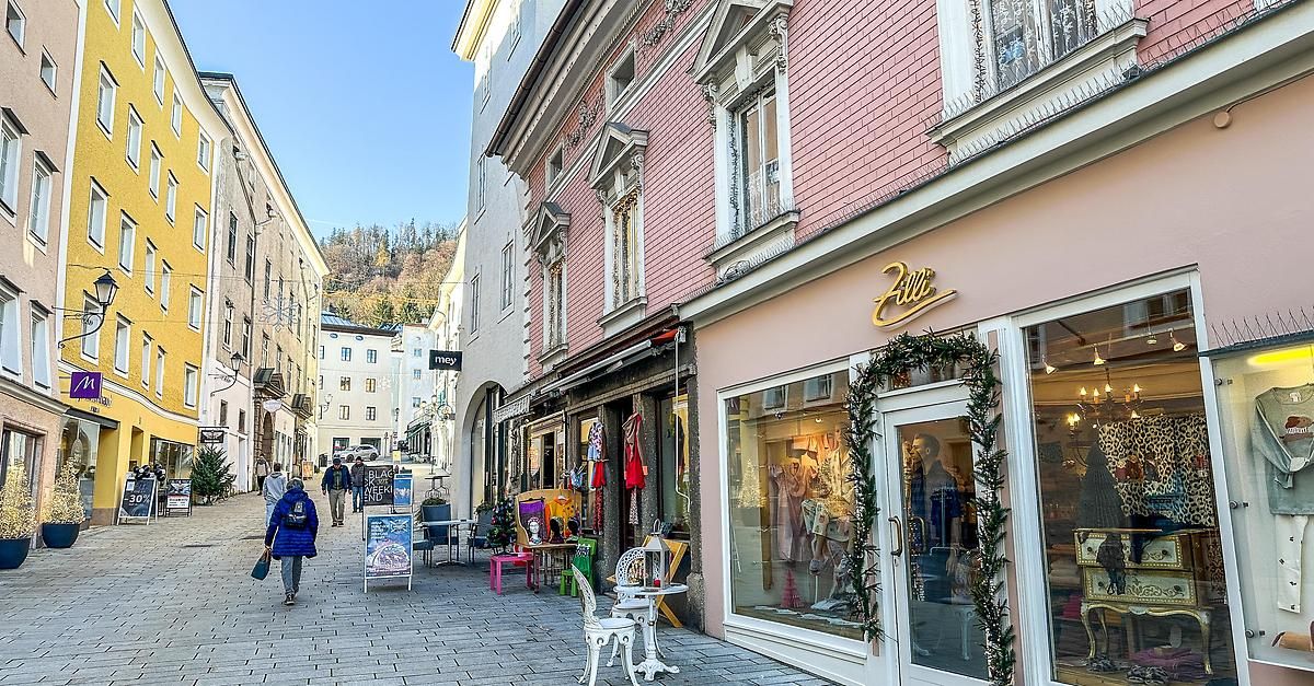 Ein Fußgänger läuft auf einer Kopfsteinpflasterstraße an Geschäften vorbei. Rechts zeigt ein Geschäft Artikel an und ist mit Grünpflanzen dekoriert. Im Hintergrund sind Gebäude und ein klarer blauer Himmel zu sehen.