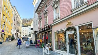 Ein Fußgänger läuft auf einer Kopfsteinpflasterstraße an Geschäften vorbei. Rechts zeigt ein Geschäft Artikel an und ist mit Grünpflanzen dekoriert. Im Hintergrund sind Gebäude und ein klarer blauer Himmel zu sehen.