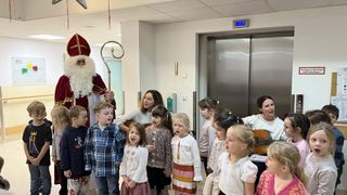 Eine Gruppe von Kindern und Erwachsenen singt in einem Flur. Ein Mann, der als Weihnachtsmann verkleidet ist, hält ein Mikrofon und steht vor ihnen. Eine Frau mit einer Gitarre steht neben ihnen. Ein Aufzug befindet sich rechts, und eine Steckdose ist an der Wand angebracht.