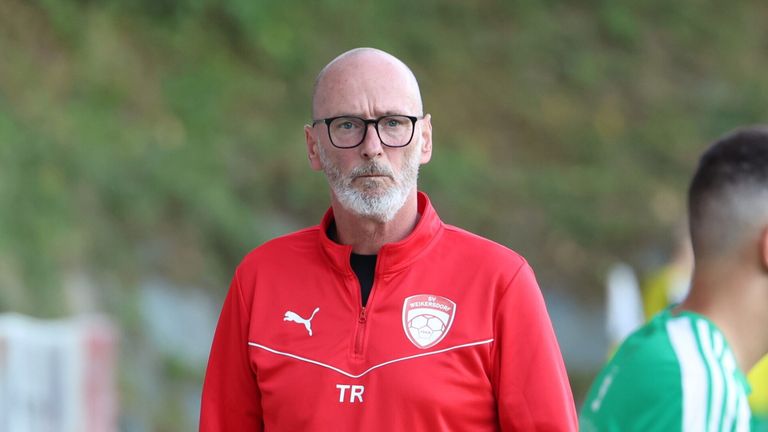 Ein Mann in einem roten Trainingsanzug mit den Buchstaben TR auf der linken Seite und einem Logo mit einem Fußball auf der rechten Seite.