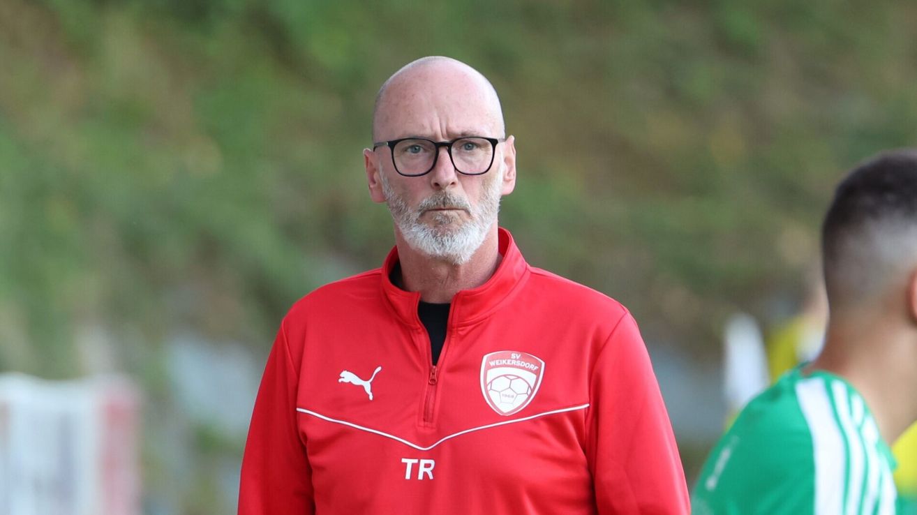 Ein Mann in einem roten Trainingsanzug mit den Buchstaben TR auf der linken Seite und einem Logo mit einem Fußball auf der rechten Seite.