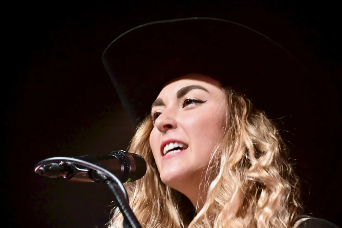 Eine Frau mit blonden Locken, die einen schwarzen Cowboyhut trägt, singt in ein Mikrofon. Sie lächelt mit leicht geöffnetem Mund. Das Mikrofon ist schwarz mit einem silbernen Griff.