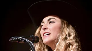 Eine Frau mit blonden Locken, die einen schwarzen Cowboyhut trägt, singt in ein Mikrofon. Sie lächelt mit leicht geöffnetem Mund. Das Mikrofon ist schwarz mit einem silbernen Griff.