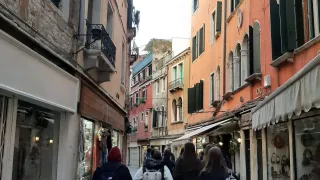 Eine enge Straße in einer Stadt mit mehreren Geschäften auf beiden Seiten, Menschen mit Rucksäcken gehen darauf.