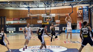 Ein Basketballspiel ist im Gange mit Spielern von zwei Teams, die aktiv beteiligt sind. Ein Spieler springt, um den Ball zu werfen, während andere sich auf dem Spielfeld bewegen. Das Spiel wird auf einem Indoor-Spielfeld mit Holzwänden und einer Anzeigetafel gespielt.