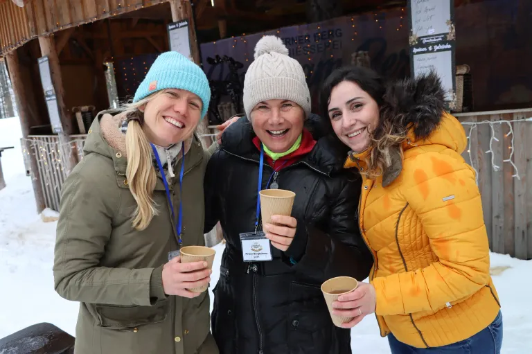 Drei Frauen in Winterkleidung, die lächeln und Tassen halten, stehen eng beieinander. Eine trägt eine schwarze Jacke, eine andere eine gelbe Jacke und die dritte eine grüne Jacke. Sie scheinen sich bei einer festlichen Veranstaltung zu befinden.