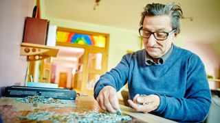 Ein älterer Mann mit Brille und blauem Pullover arbeitet an einem Puzzle auf einem Tisch. Der Tisch ist mit Puzzleteilen bedeckt. Dahinter befindet sich eine Holztür und ein Buntglasfenster.