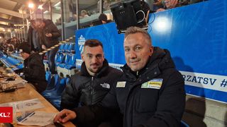 Zwei Männer sitzen auf einer Bank bei einer Sportveranstaltung. Sie tragen schwarze Jacken und lächeln für ein Foto. Der Mann auf der linken Seite hat ein 'The North Face' Logo. Der Hintergrund besteht aus leeren blauen Stühlen und einer blauen Barriere.