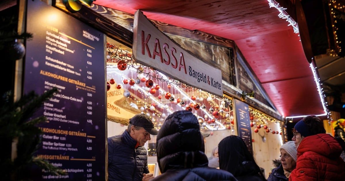 Ein Mann spricht mit einem Verkäufer an einem KASSa-Stand auf einem Weihnachtsmarkt. Der Stand hat ein Schild über ihm mit 'KASSa' in Rot und 'Bargeld & Karte' in Schwarz. Dekorationen hängen von der Decke.