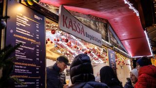 Ein Mann spricht mit einem Verkäufer an einem KASSa-Stand auf einem Weihnachtsmarkt. Der Stand hat ein Schild über ihm mit 'KASSa' in Rot und 'Bargeld & Karte' in Schwarz. Dekorationen hängen von der Decke.