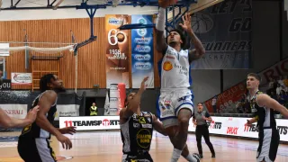 Basketballspieler spielen auf einem Hallenplatz. Ein Spieler in einem weißen Trikot springt und versucht, den Ball zu werfen. Ein anderer Spieler in einem schwarzen Trikot verteidigt.