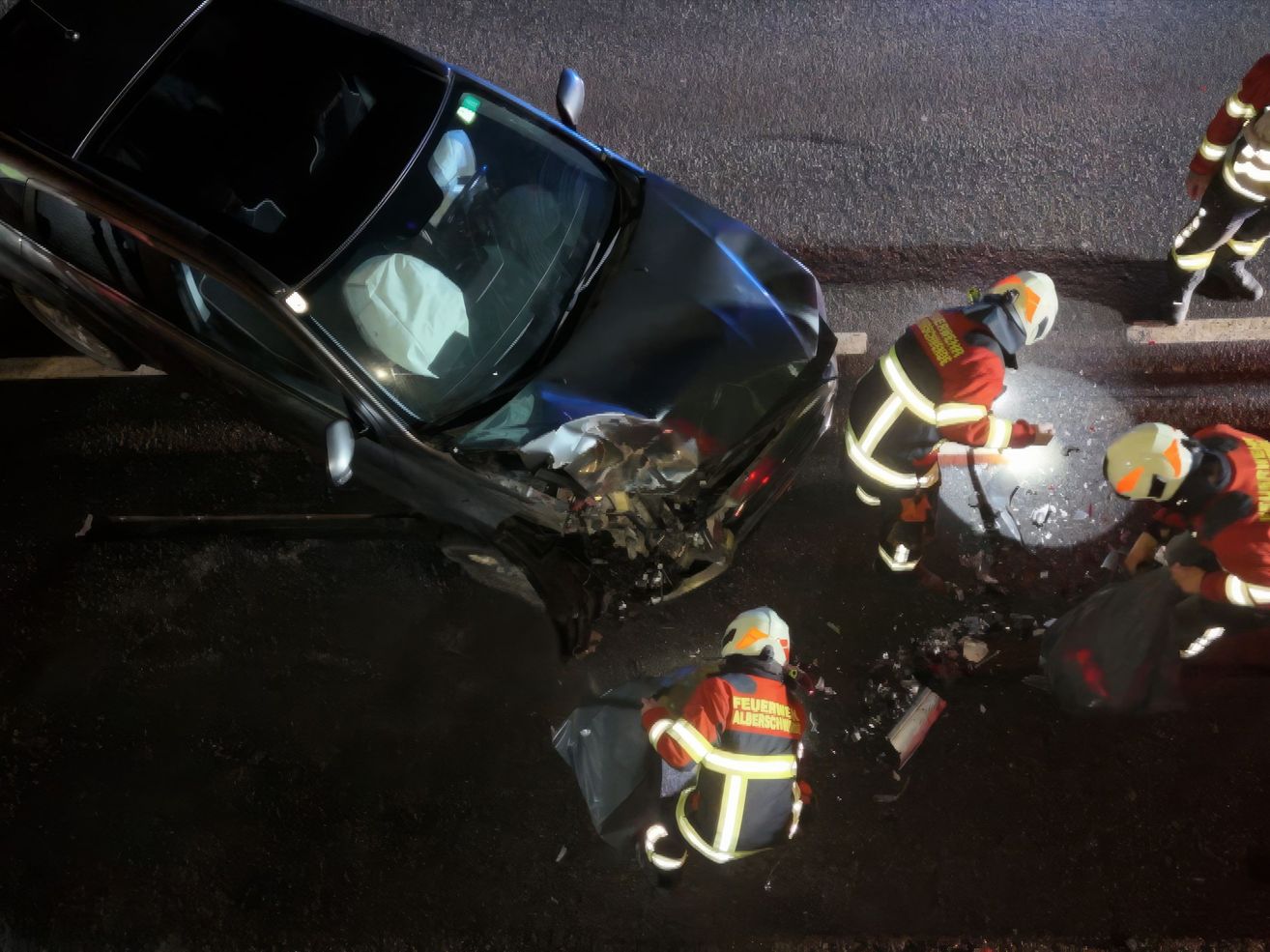 Feuerwehrleute kümmern sich um einen Autounfall. Ein Auto hat erhebliche Frontschäden. Zwei Feuerwehrleute sind am Unfallort, einer trägt einen Helm und hält ein Werkzeug.