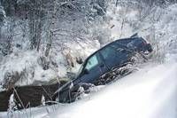 Ein dunkelblaues Auto steckt im Schnee in der Nähe eines Waldes fest, teilweise auf der Seite eingesunken. Das Auto ist schneebedeckt, und das Gebiet ist von Bäumen umgeben.