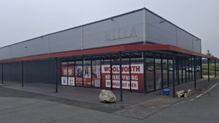 Ein Gebäude mit rotem Vordach hat ein großes Schild mit der Aufschrift Woolworth. Das Schild zeigt Bilder von Menschen und einen QR-Code. Ein großer Stein liegt auf dem Bürgersteig.