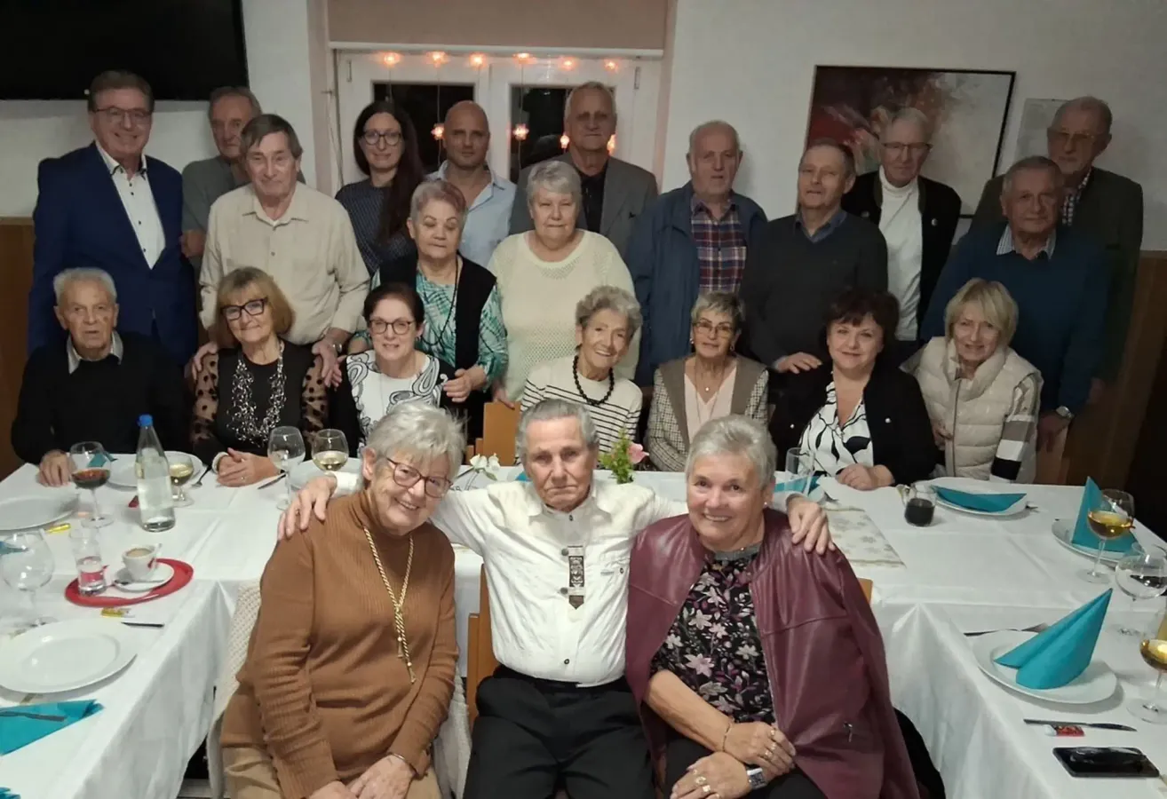 Eine große Gruppe älterer Personen posiert für ein Foto in einem gut beleuchteten Raum. Einige sitzen an einem Tisch, während andere dahinter stehen. Sie alle lächeln für die Kamera.