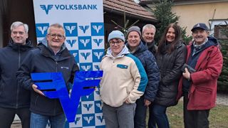 Fünf Personen stehen vor einem Volksbank-Banner mit einem Weihnachtsbaum im Hintergrund. Ein Mann hält einen blauen F-Buchstaben, während andere lächeln und glücklich aussehen.